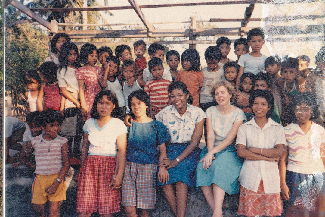 Jacqueline Huggins, seated at the center of the front bottom row in 1987, surrounded by children, illustrating the idea that children are the best language teachers.